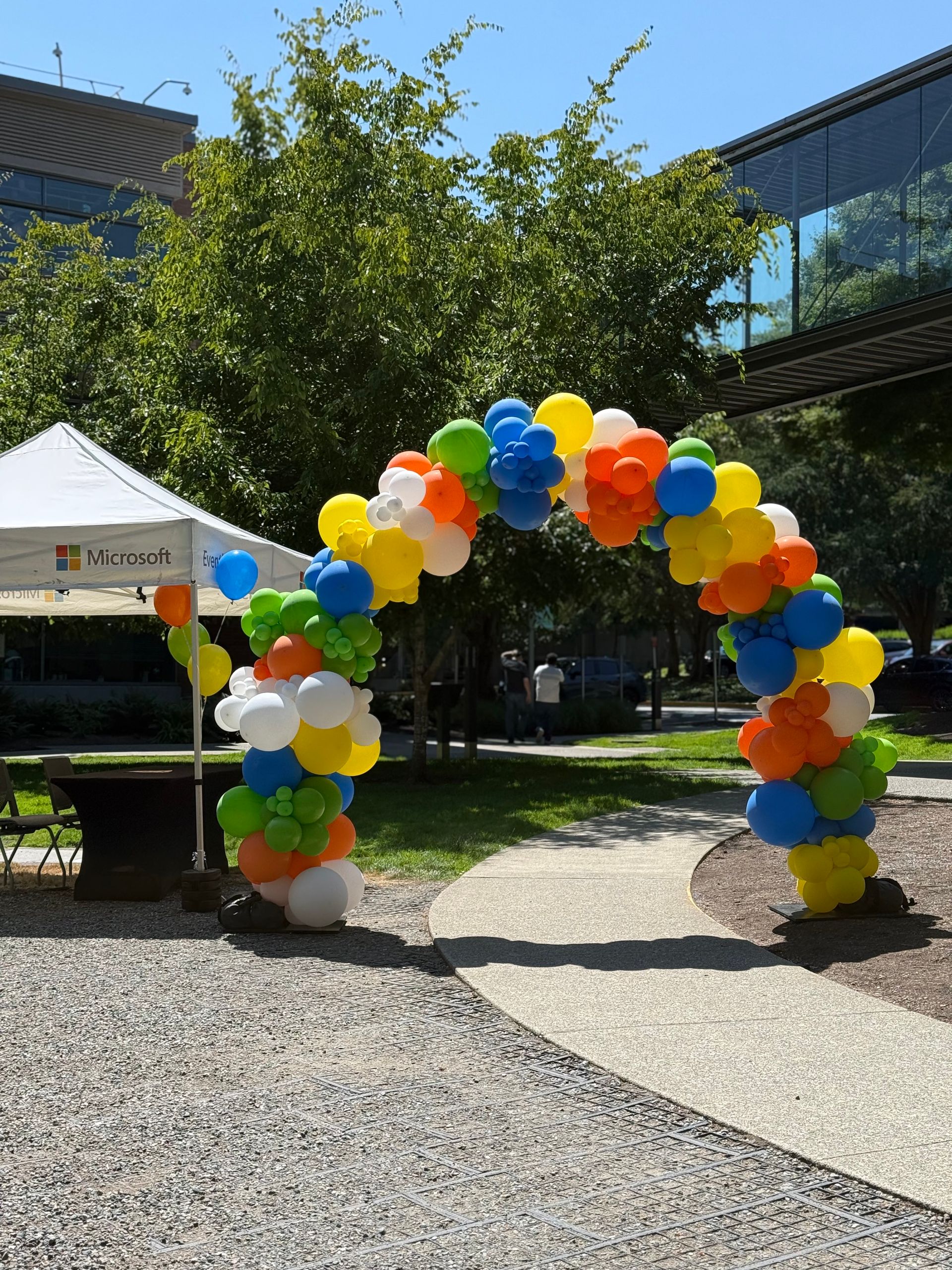 Colorful balloon arch over a gravel path outdoors beside a white tent and modern building