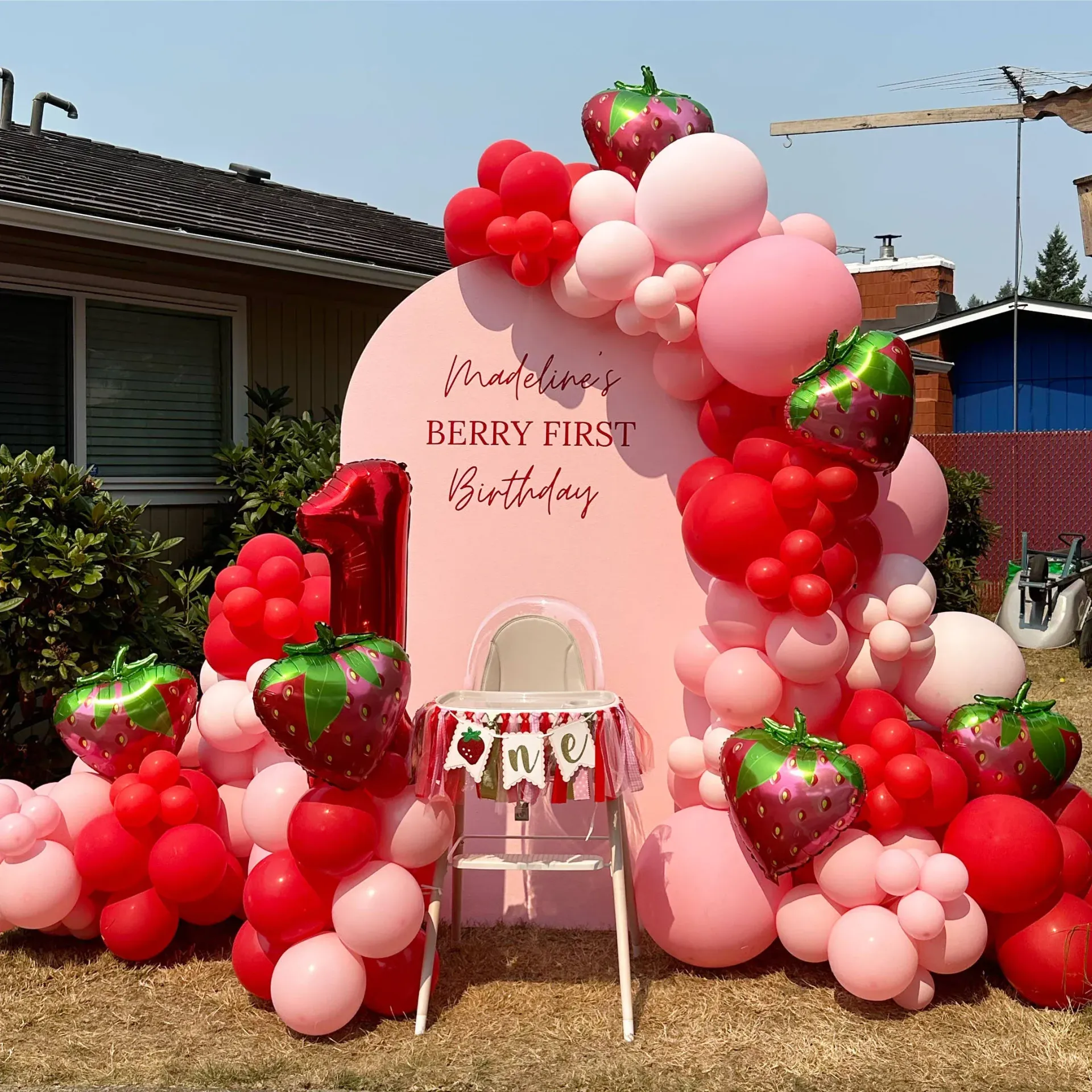 A chair is surrounded by balloons and strawberries in front of a house.