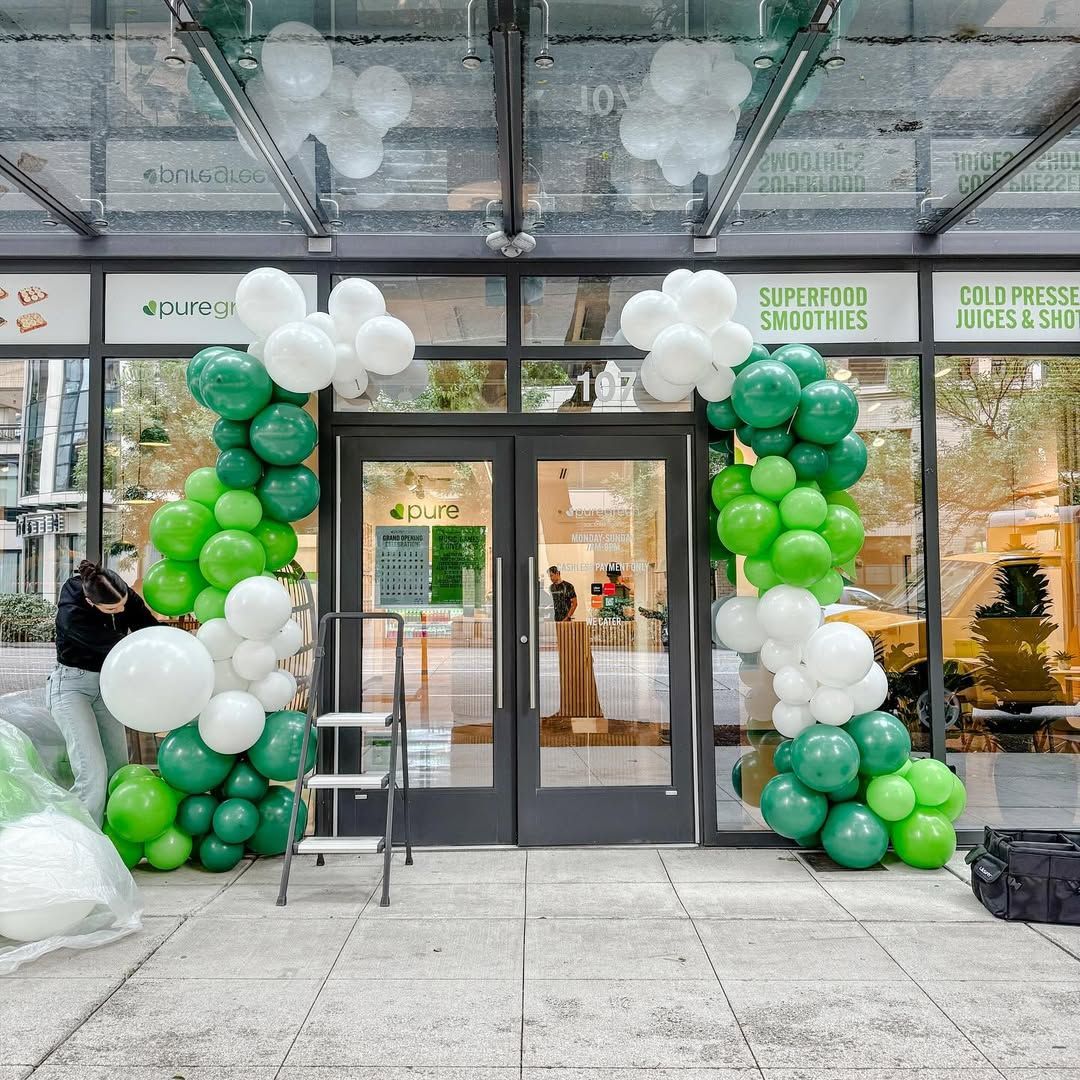 Green and white balloons decorate a store entrance. A person works on the balloons, setting them in an arch.