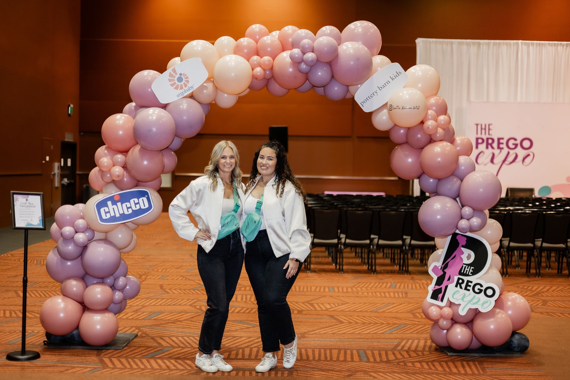 Two women are standing under a balloon arch in a room.