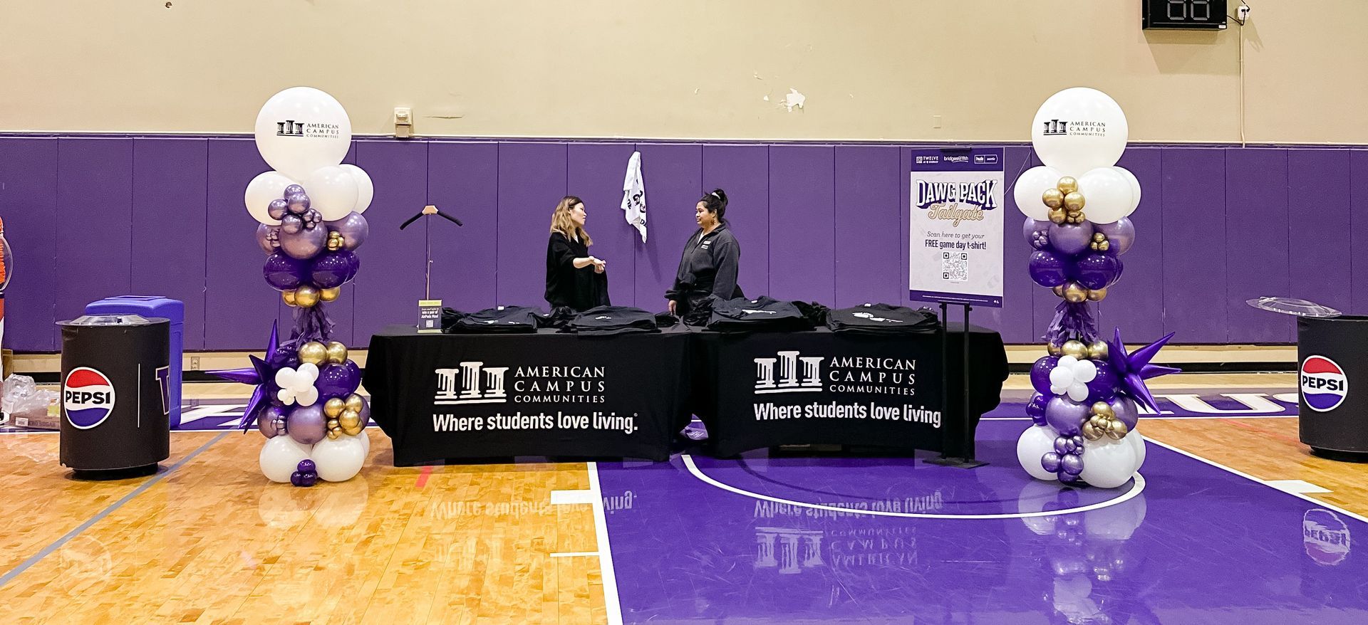 People at a table inside a gym. Balloons and banners on display, and purple and gold decorations.