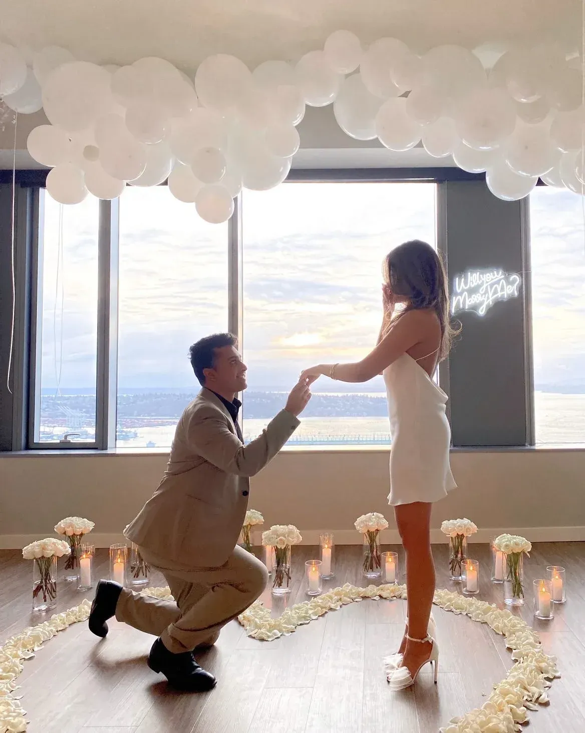 A man is kneeling down to propose to a woman in front of a window.