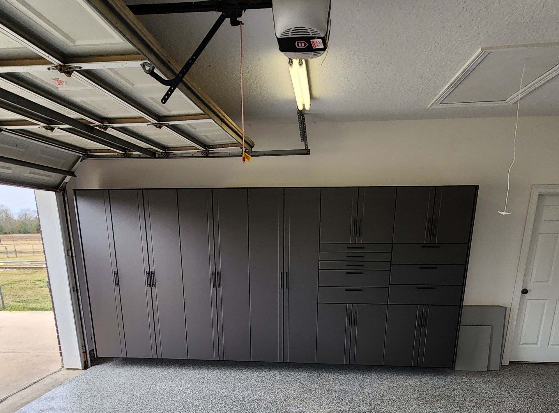 Black cabinet wall in a garage, with overhead opener, fluorescent light, and gravel floor nearby