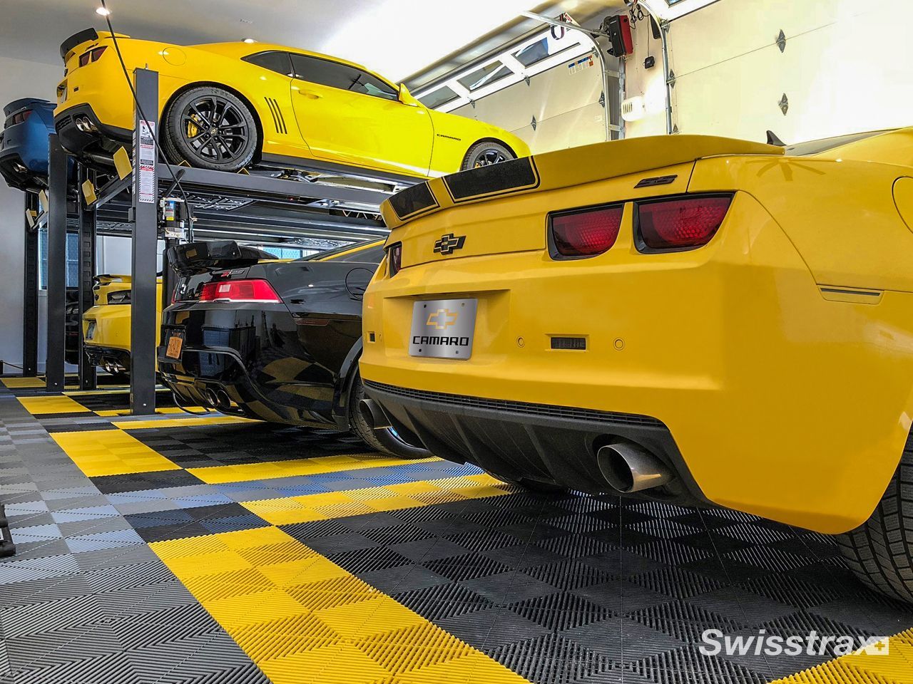 Two yellow sports cars in a stacked parking garage, one on an upper lift and one below.