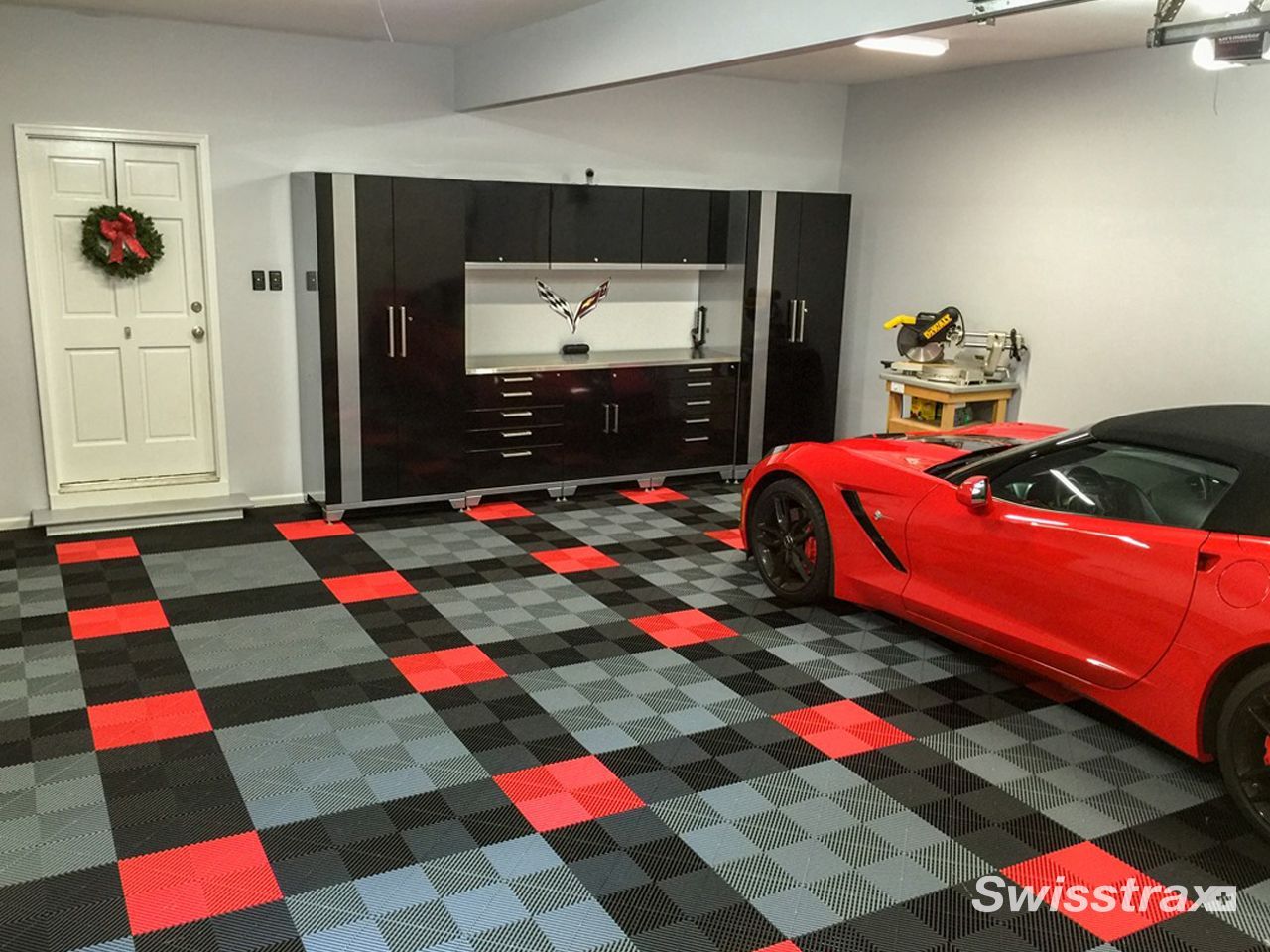 Red sports car in a garage with black, red, and gray checkered floor tiles and a dark cabinet wall.