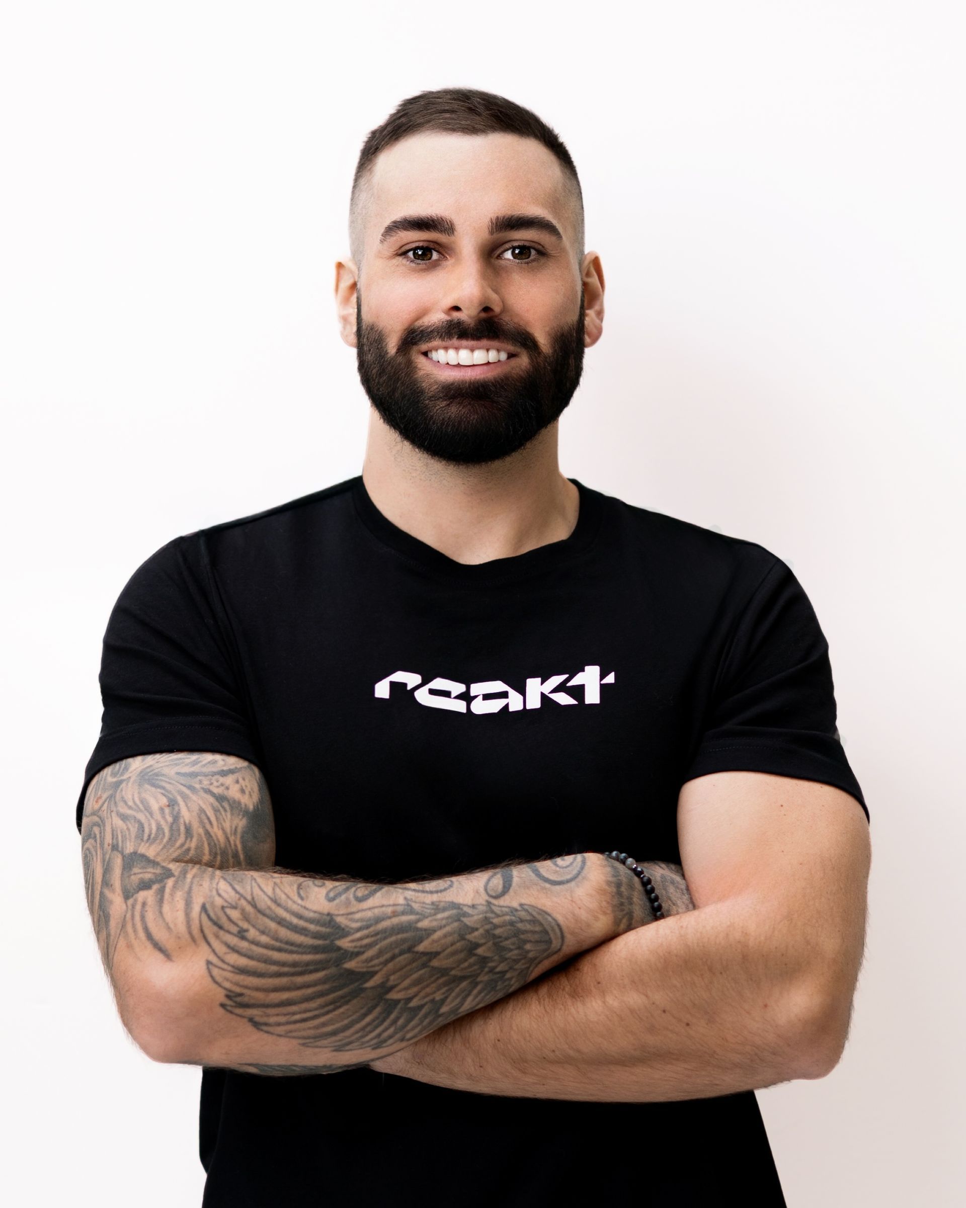 A smiling person with a beard and tattoos on their arms stands with arms crossed against a plain, light background.