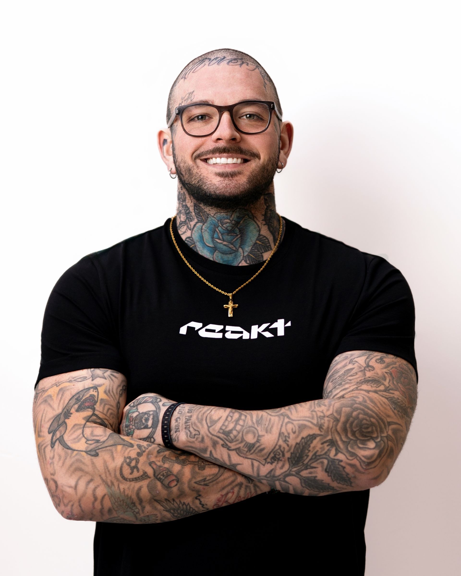 A tattooed person with glasses and a cross necklace smiles with arms crossed, wearing a black t-shirt against a white wall.