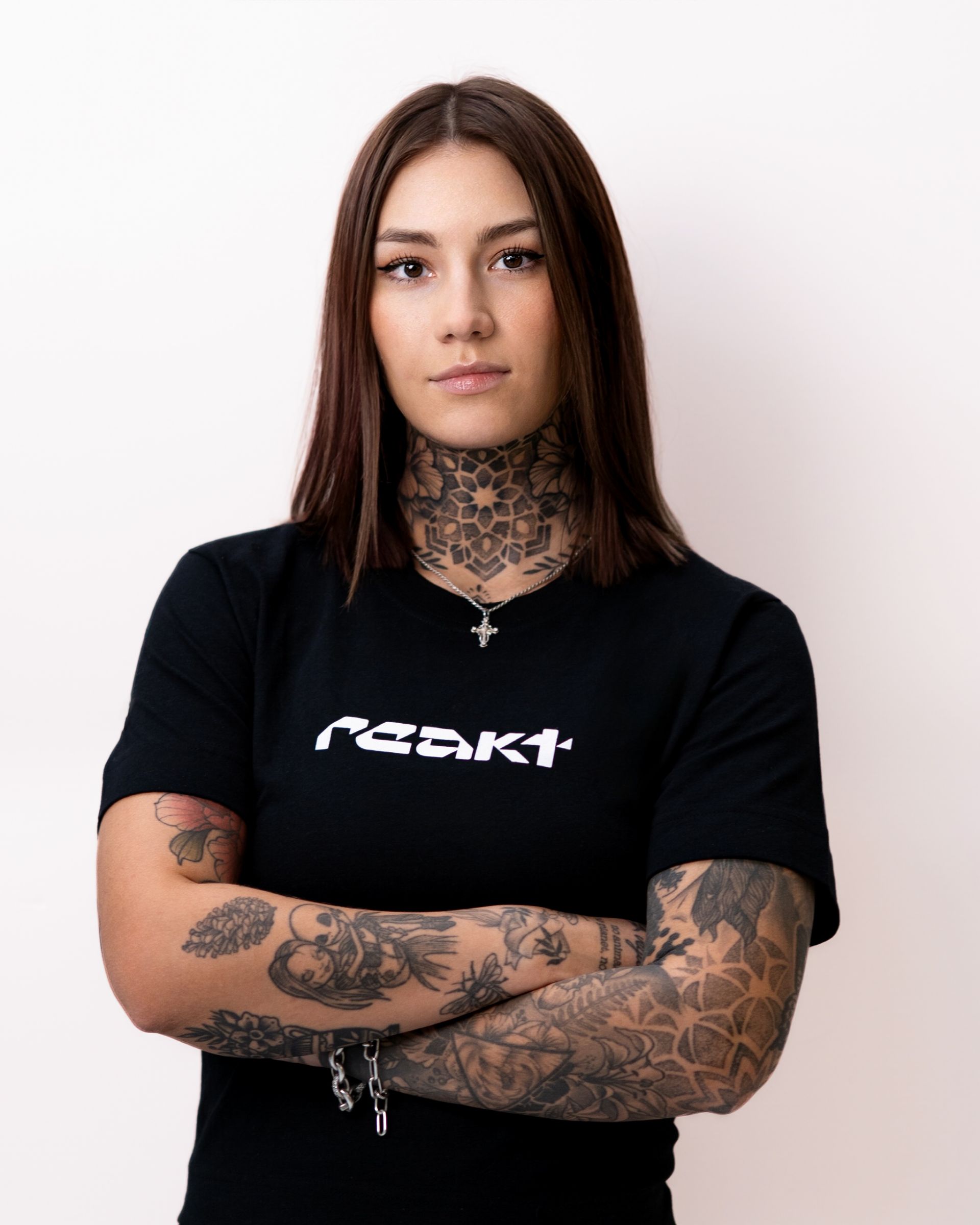 A person with extensive arm and neck tattoos stands against a white background, arms crossed, wearing a black logo t-shirt.