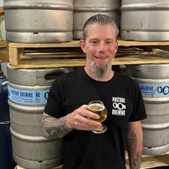 a man with a beard is holding a glass of beer in a brewery