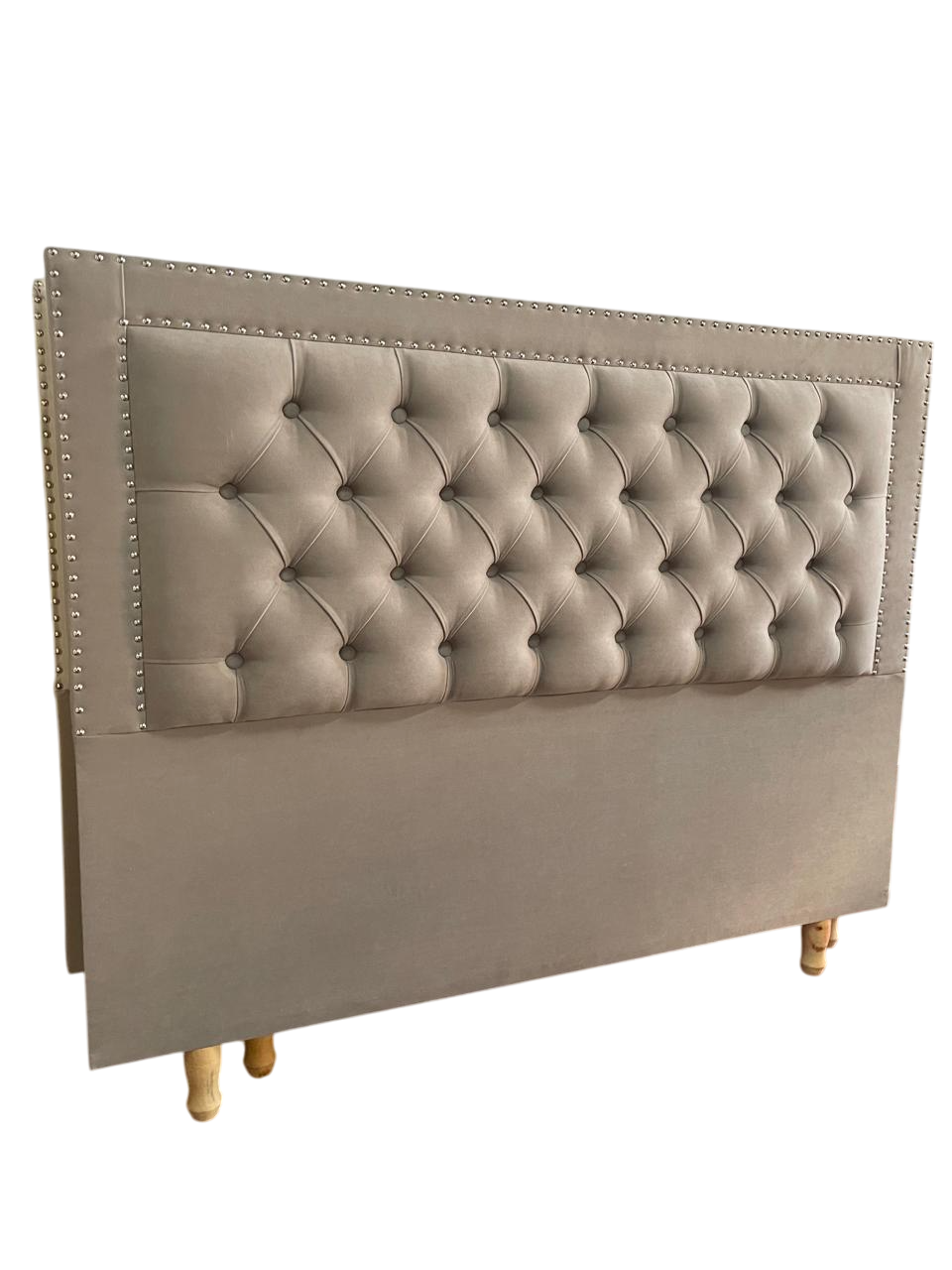 Cabecero capitoné beige con ribete de tachuelas sobre patas de madera.