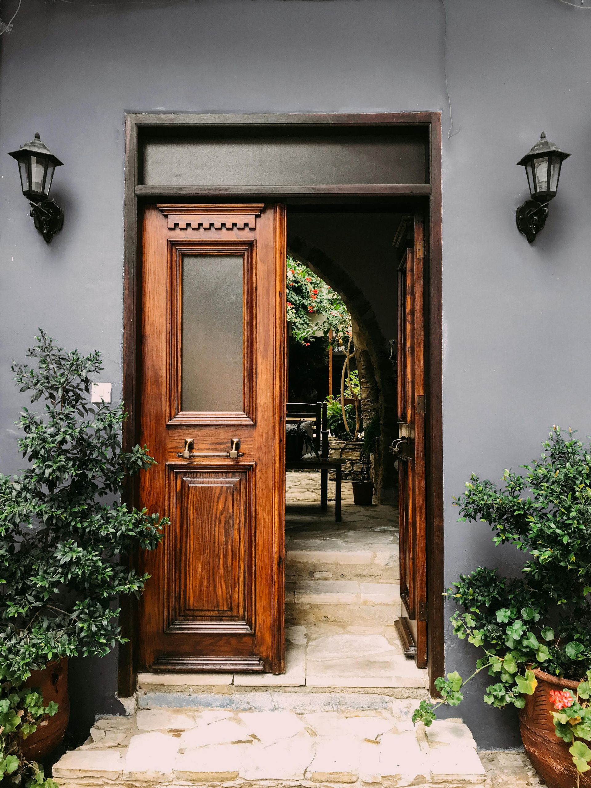 Wooden door, ajar, reveals a stone archway leading to greenery. Gray walls, potted plants flank the entrance.