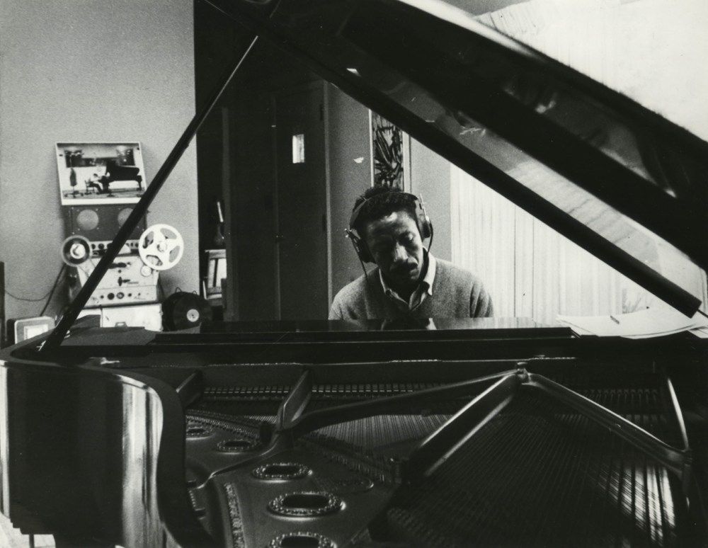 (Gordon Parks Foundation) Parks sits at a piano with headphones on. We are looking at him from the foot of the piano, through the open top of the baby grand, the strings pulled tight inside the instrument. He is focused on the keys before him and seems to not know that the photo is being taken. 
