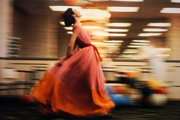 A woman stands in a floor-length pink and orange, chiffon dress.  the dress is belted with a reddish orange sash and the long skirt of the dress billows behind her as she throws her arms and shoulders back and smiles toward the ceiling. The photo, other than the womans face, seems to be caught in the middle of a movement and therefore is a blur behind her. She appears to be in a department store