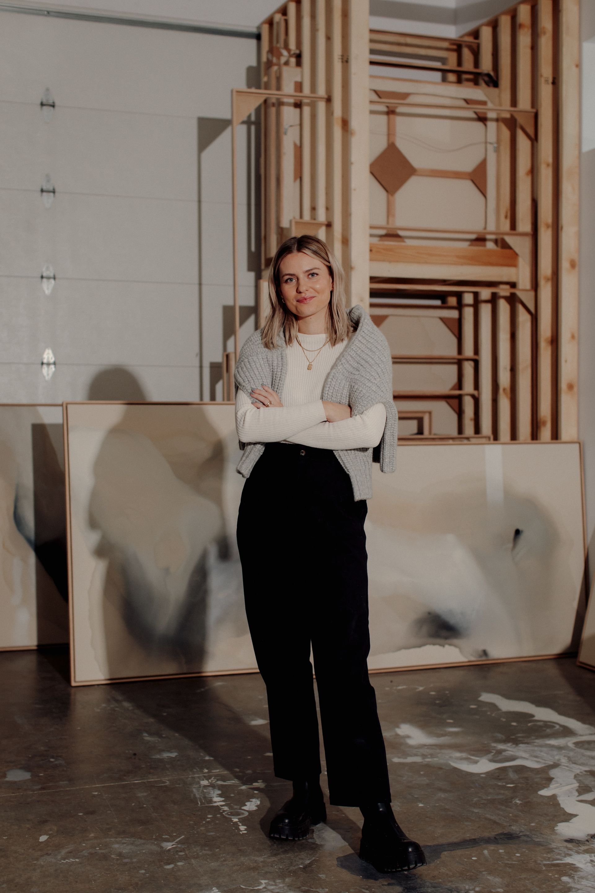 Joy Kinna in her studio standing in front of a stack of paintings with her arms crossed over her chest and a sweater over her shoulders.  shot by Brit Gill Photography