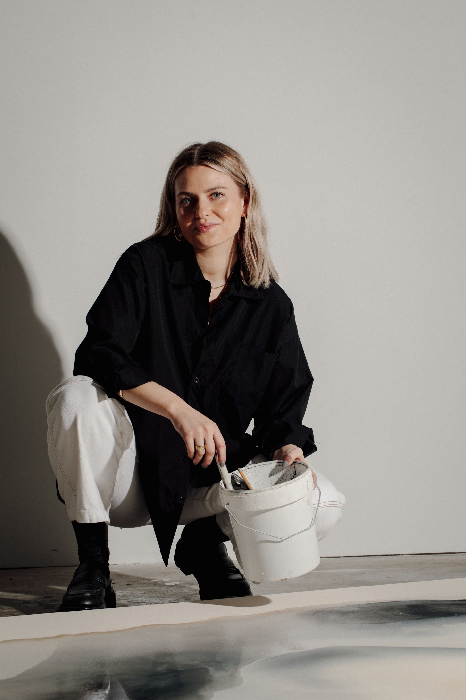 Joy Kinna  crouching over a painting as she works on it. She looks into the camera and smiles with paintbrush and water bucket in hand. shot by Brit Gill Photography