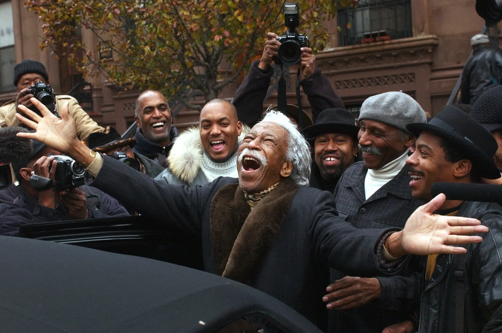 Parks celebrates with fellow photographers following a portrait session featuring himself and 100 other prominent African-American photographers in Harlem, New York, 2002.  Photograph by Suzanne Plunkett / Associated Press. Parks stands with his arms outstretched and joy erupting from his face. He is surrounded by happy looking men, all smiling at him, and a few taking his photo. 