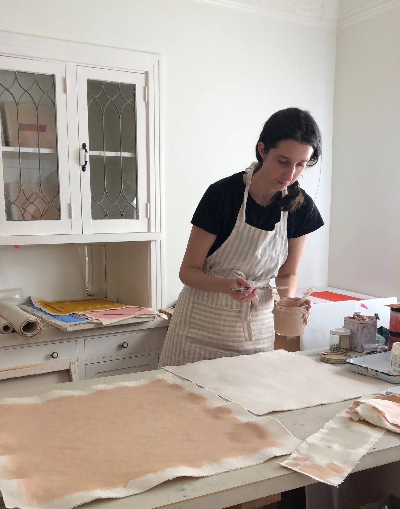 Emily Keating Snyder in her studio painting loose canvas with a peachy pink color. She is wearing an apron and leans over the canvas with brush in hand 