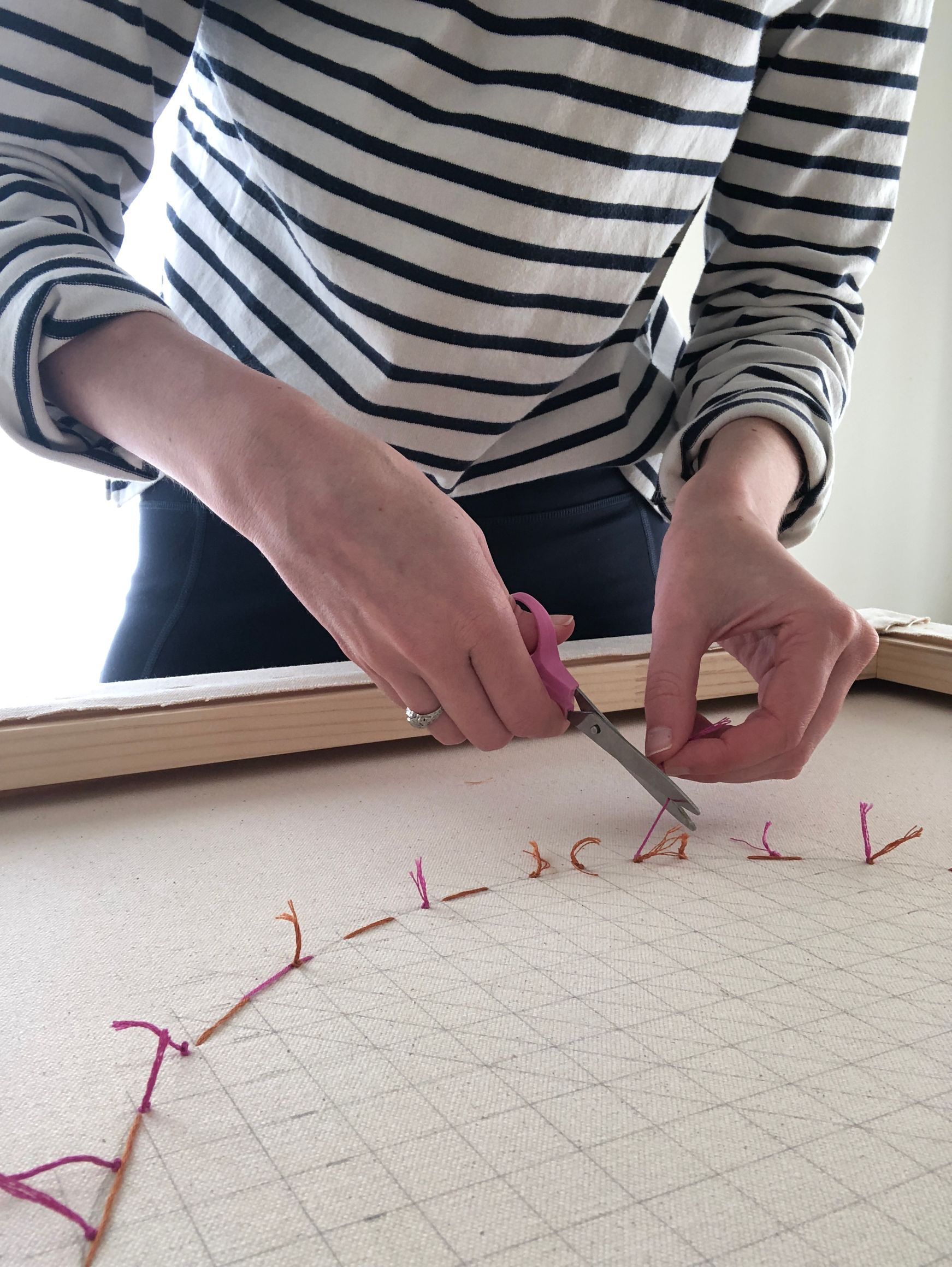 Emily Keating Snyder in her studio cutting the lose edges on the back of the piece after she is finished embroidering the front. The image is a close up of her hands with a pair of scissors in one hand and loose embroidery floss in the other 