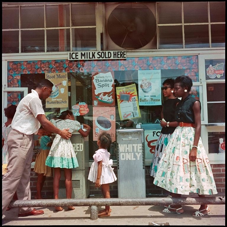 Three adults and four children stand outside a colorful ice cream shop and wait in line to drink from the 