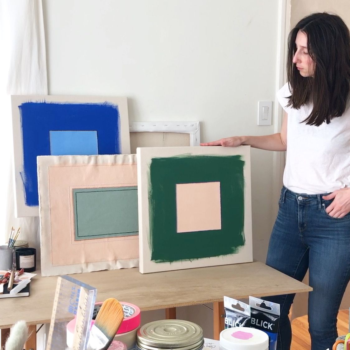 Emily Keating Snyder in her studio. She looks at a green piece of art next to her on the table stacked against a pink piece and a blue piece. 