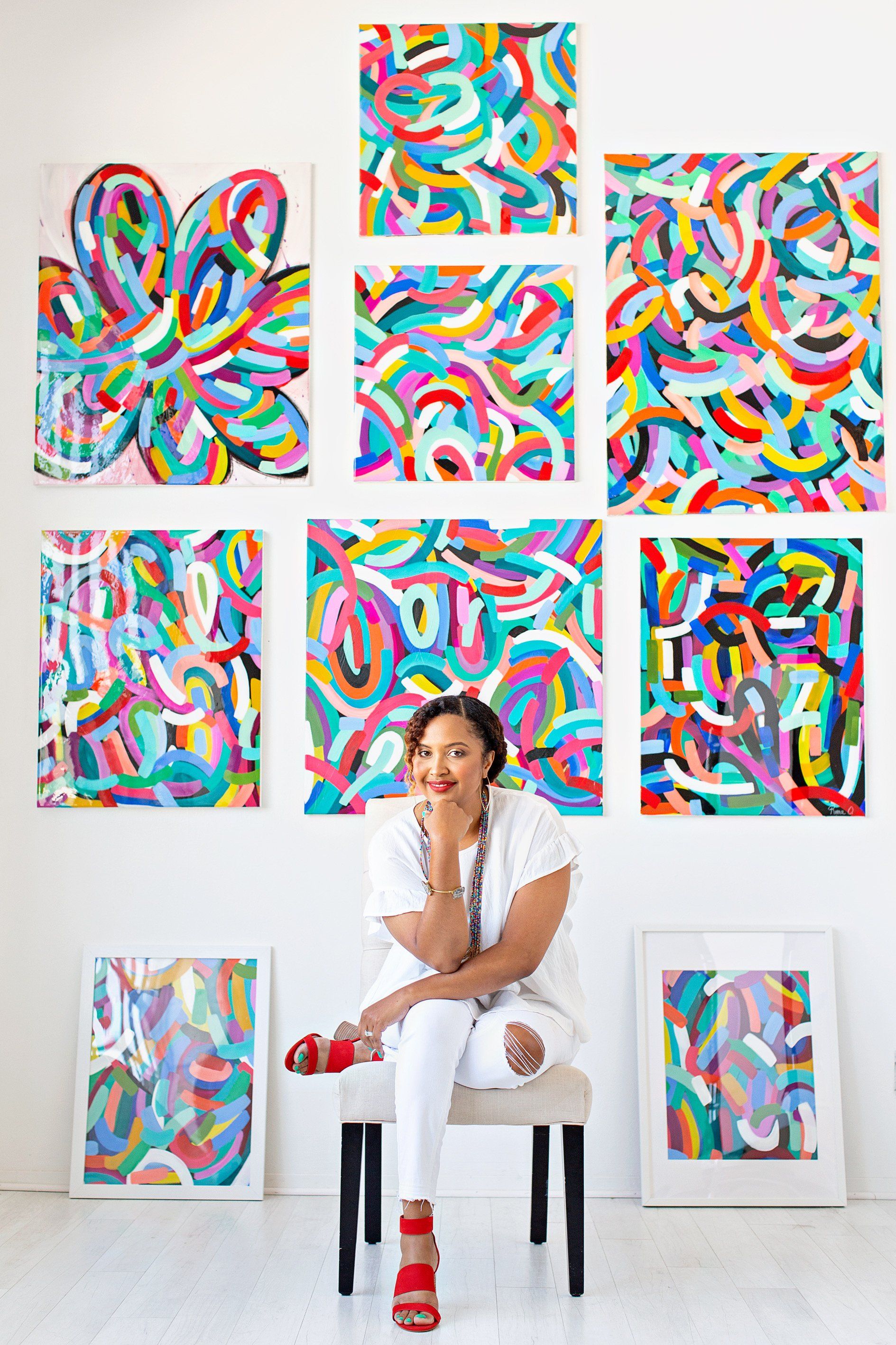 Roma Osowo poses with her artwork on the wall behind her as she rests her elbow on her knee and her chin on her elbow. 