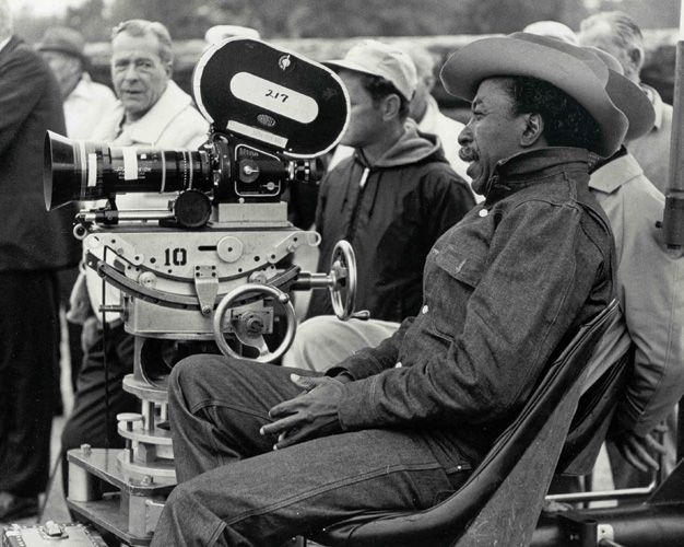 Parks sits in his chair with a large smile on his face. We see the side profile of him with the movie crew behind him, filming, with the large camera next to him. He is wearing a denim top and pants with a small cowboy hat on his head. 