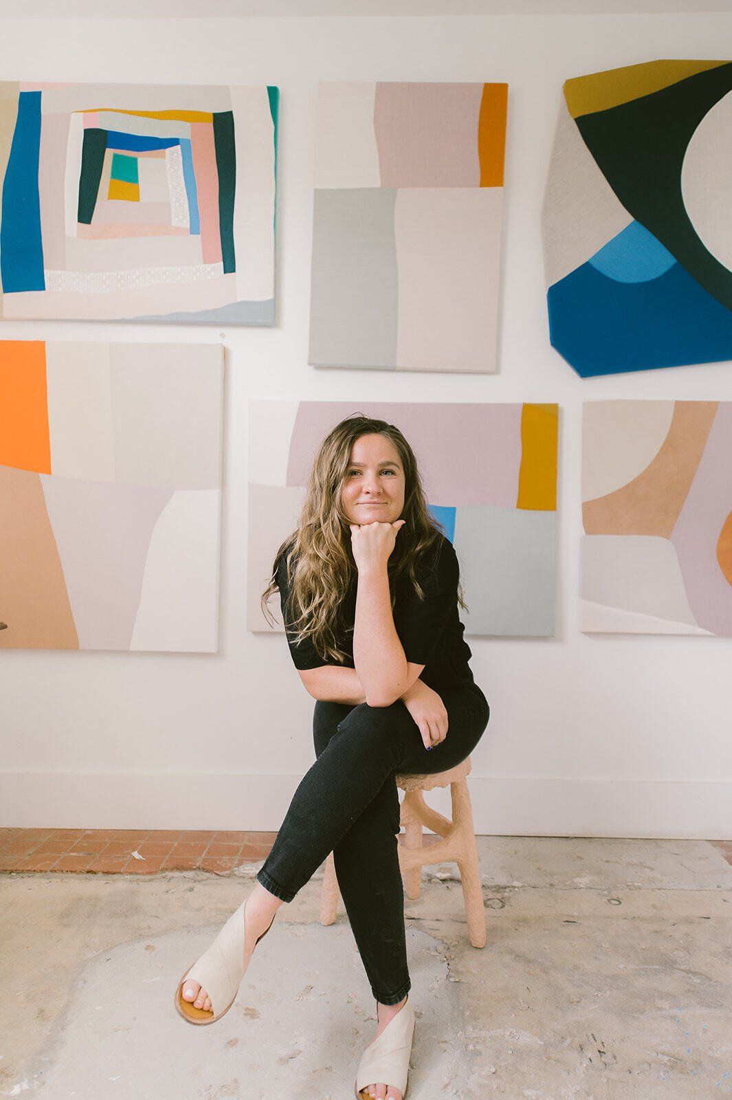 Sydney in her studio with art behind her. she is sitting on a stool, resting her elbow on her  crossed legs and propping her head up, smiling softly. 