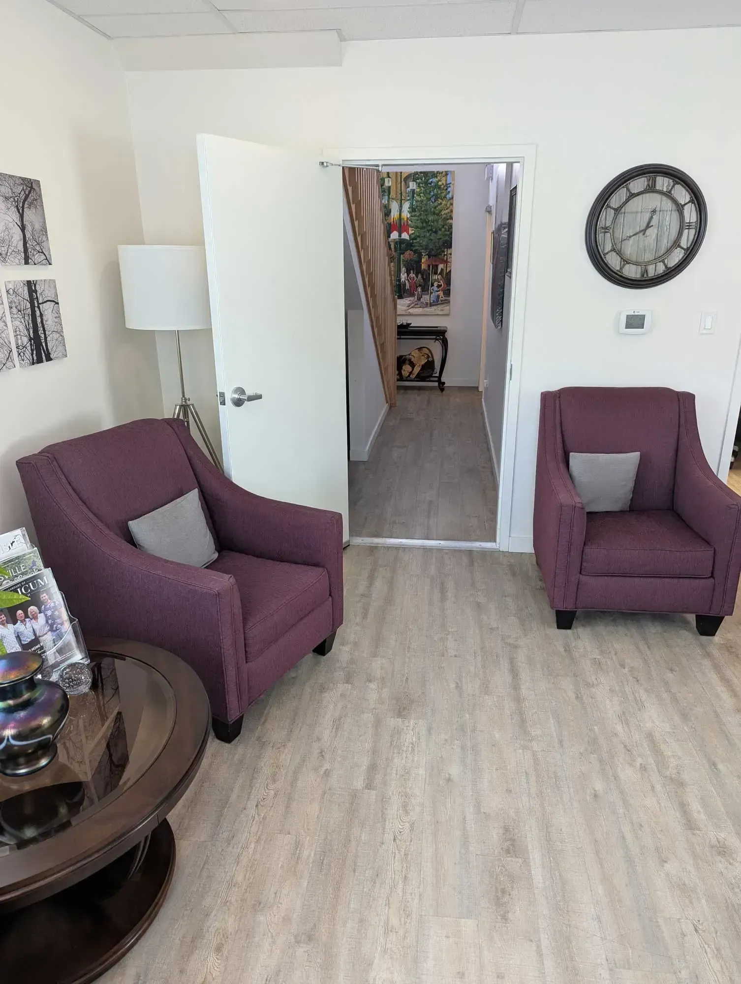 Two purple armchairs in a waiting area with a round table, clock, and doorway to a staircase.