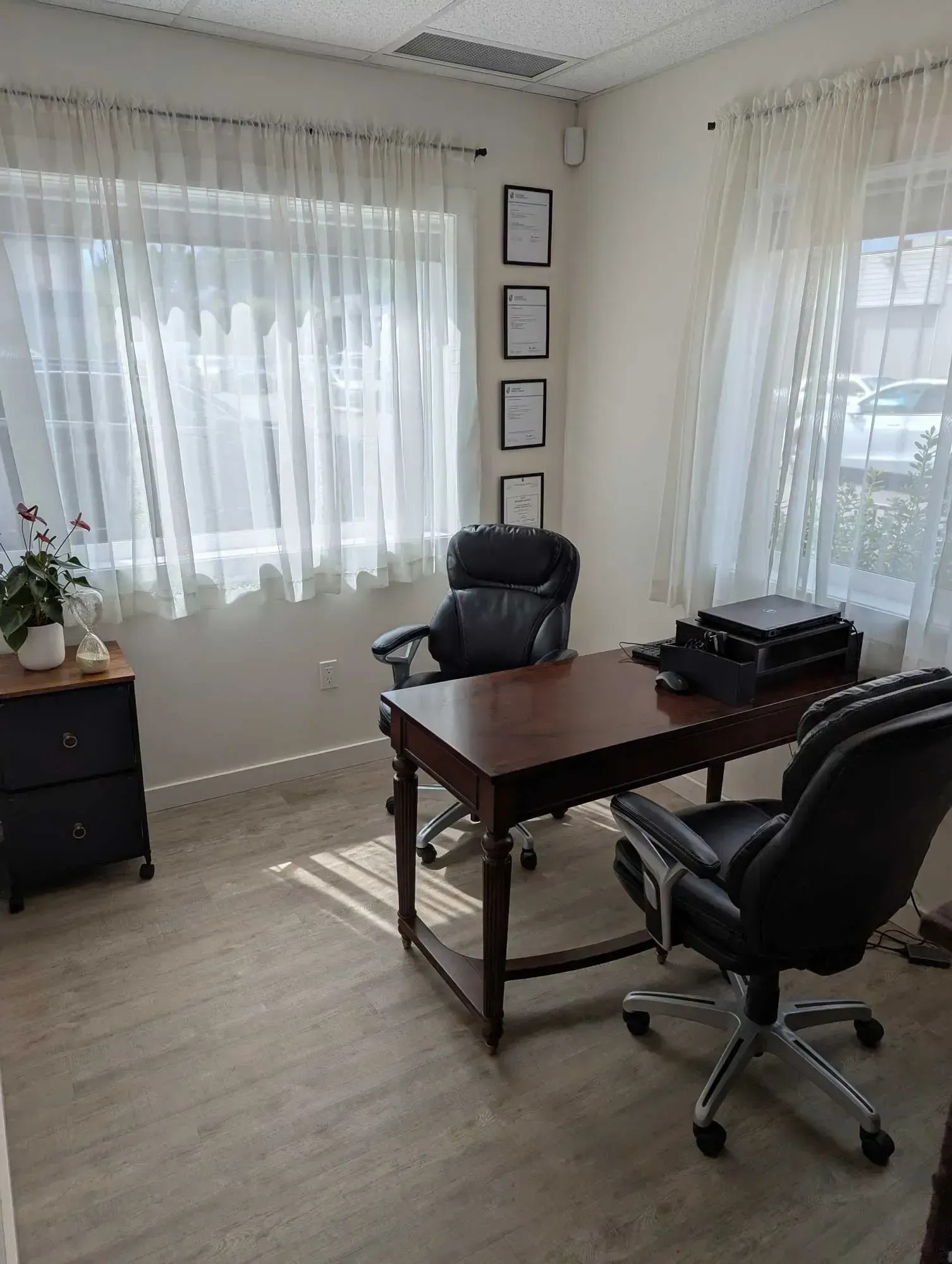 Office with a desk, two black chairs, windows with sheer curtains, and framed documents on the wall.