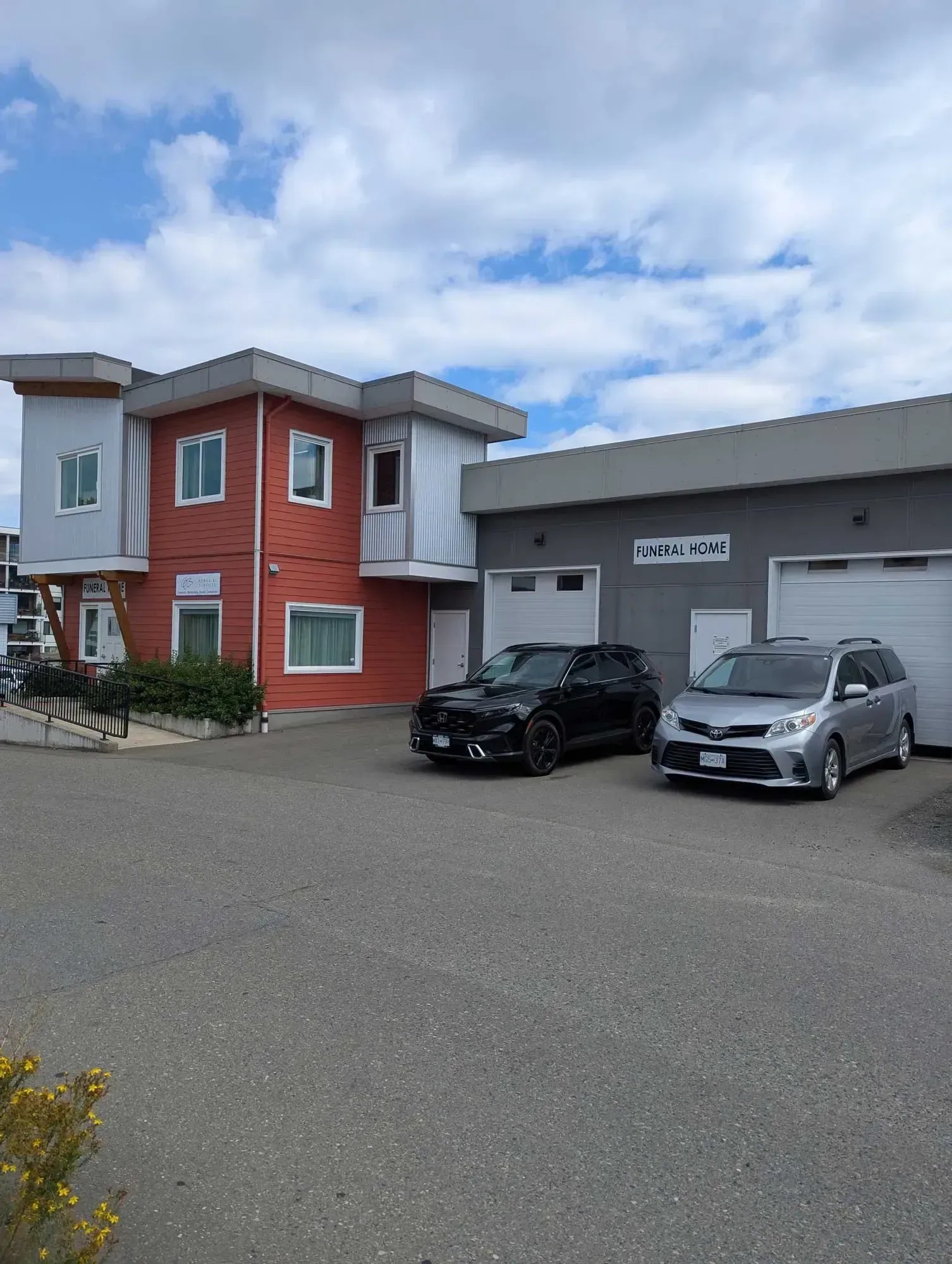 A two-story red brick building with a black SUV and silver minivan parked in front. Gray building with garage doors. Blue sky.