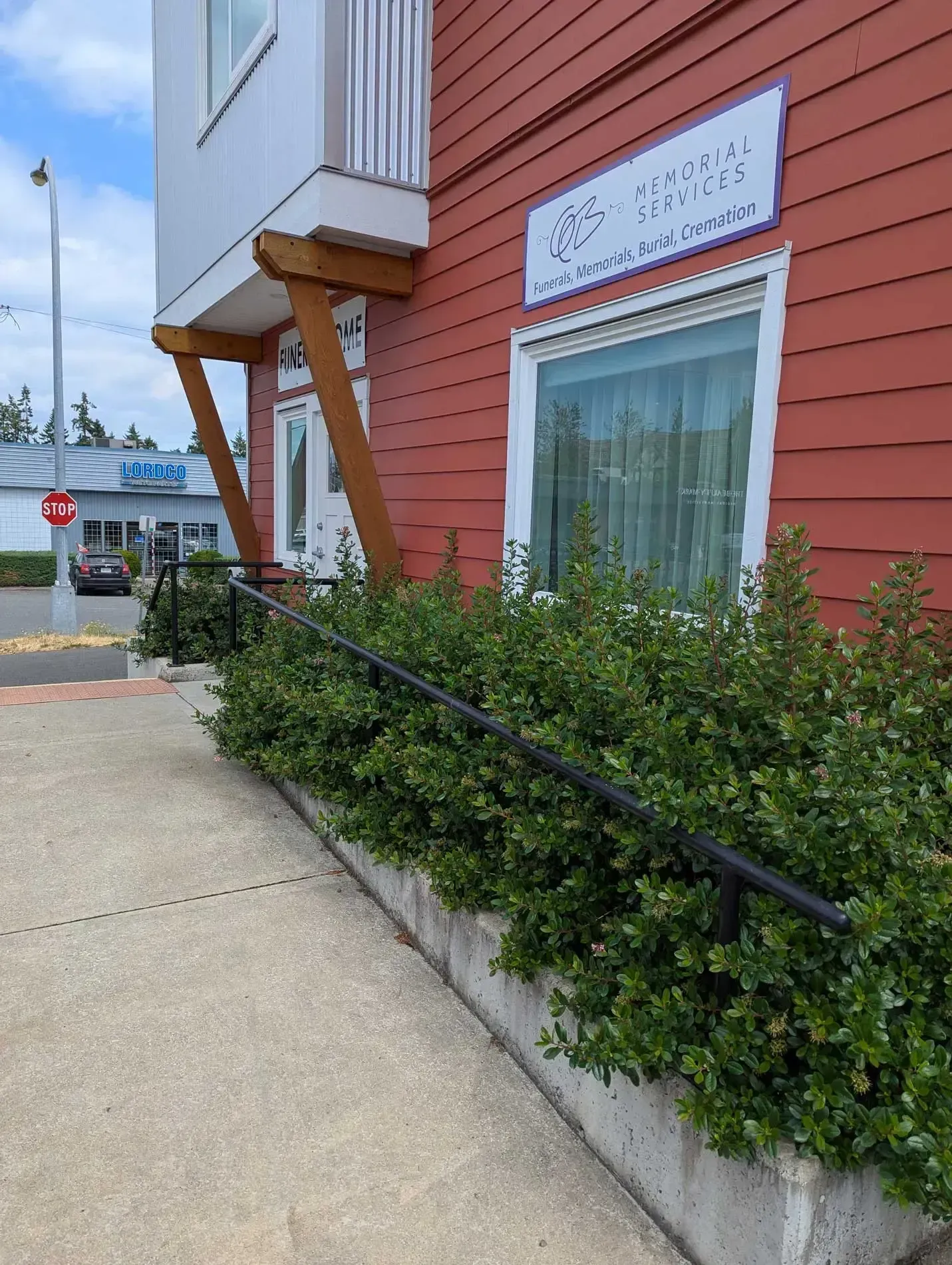 Red building with a sign and a ramp with a black handrail and a row of green bushes.
