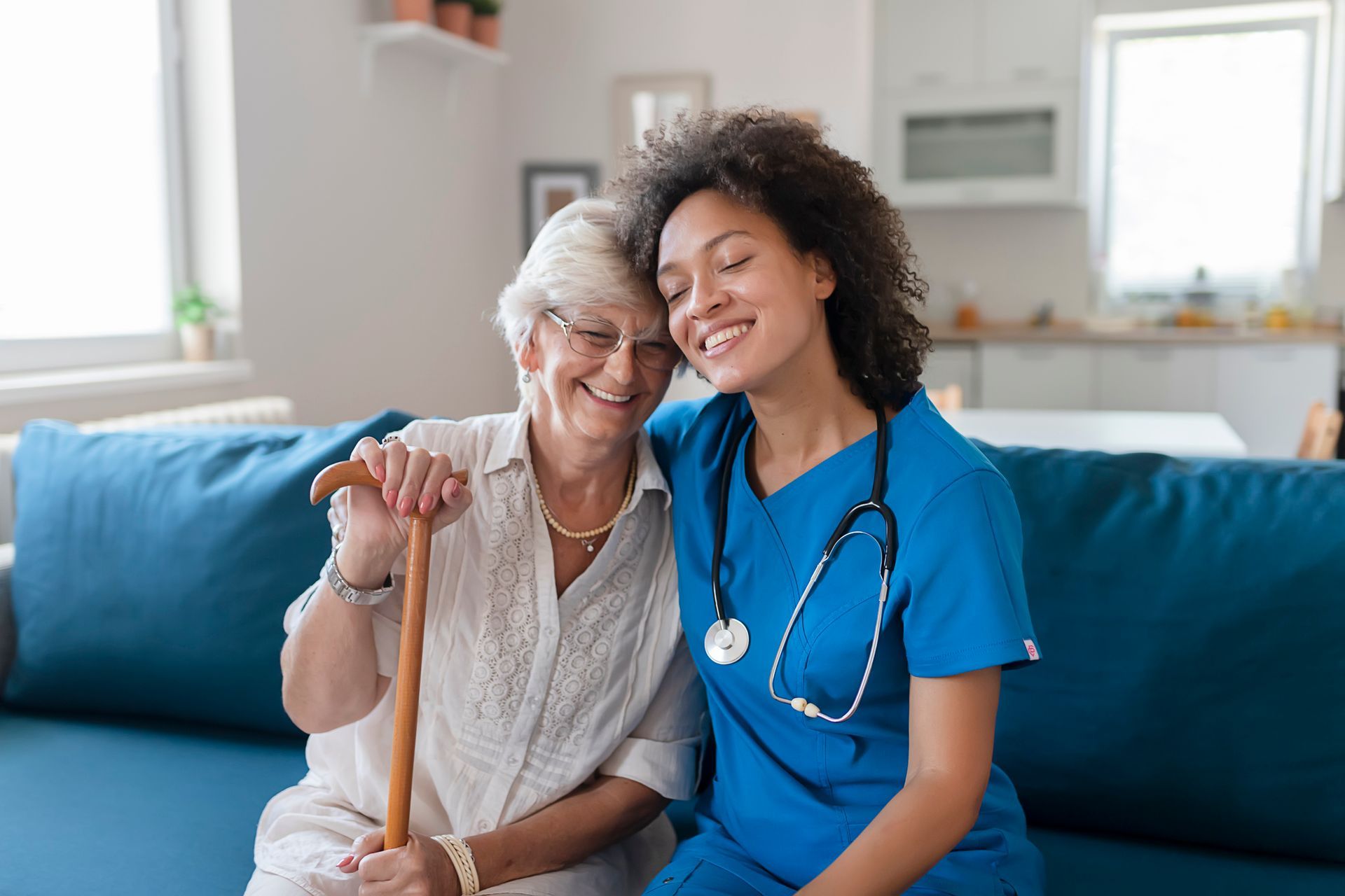A nurse is sitting on a couch with an elderly woman holding a cane.