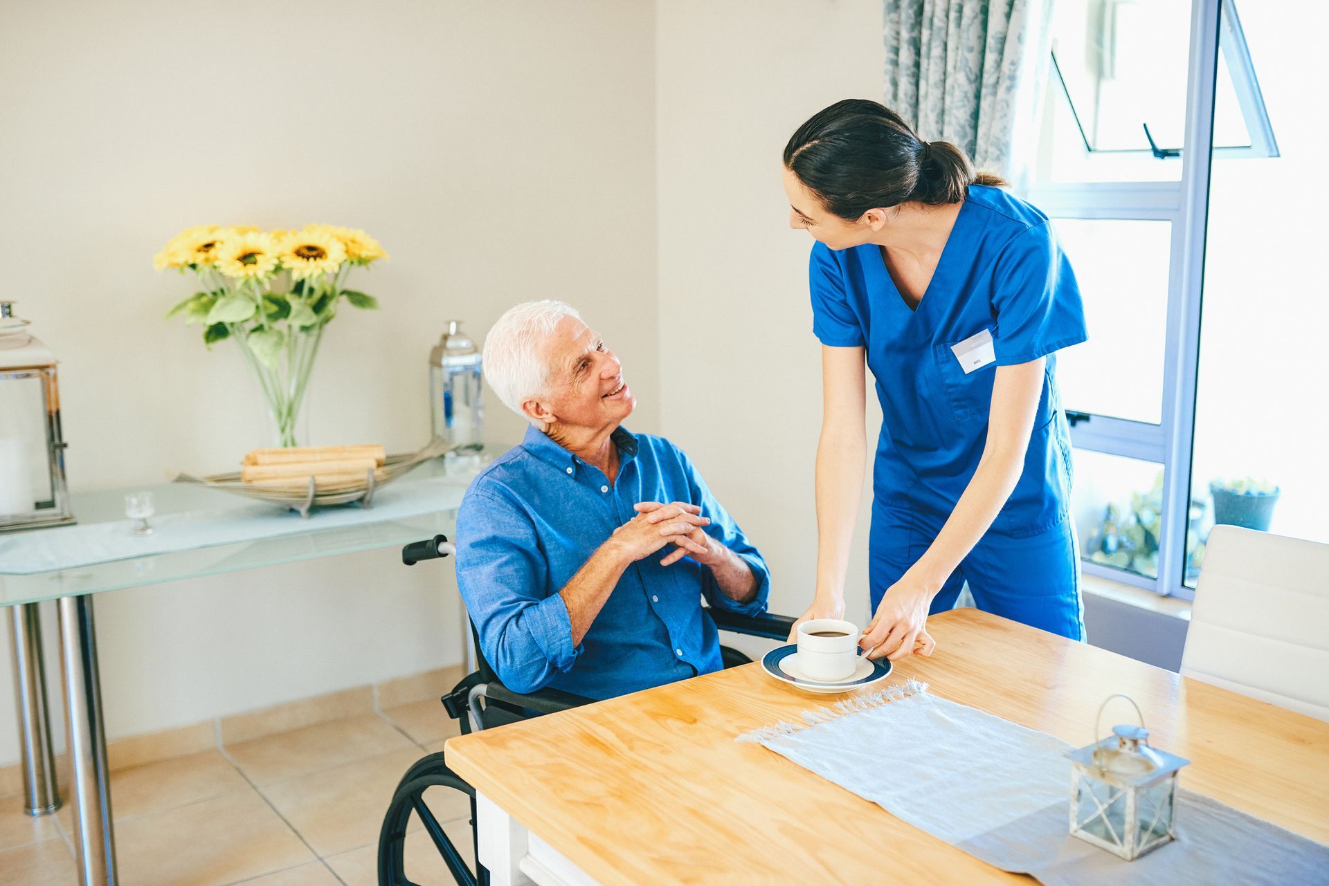 A nurse is serving a cup of coffee to an elderly woman in a wheelchair.