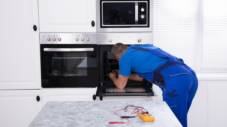 A technician in blue workwear repairs an oven in a white kitchen, with a multimeter placed on a marble counter.