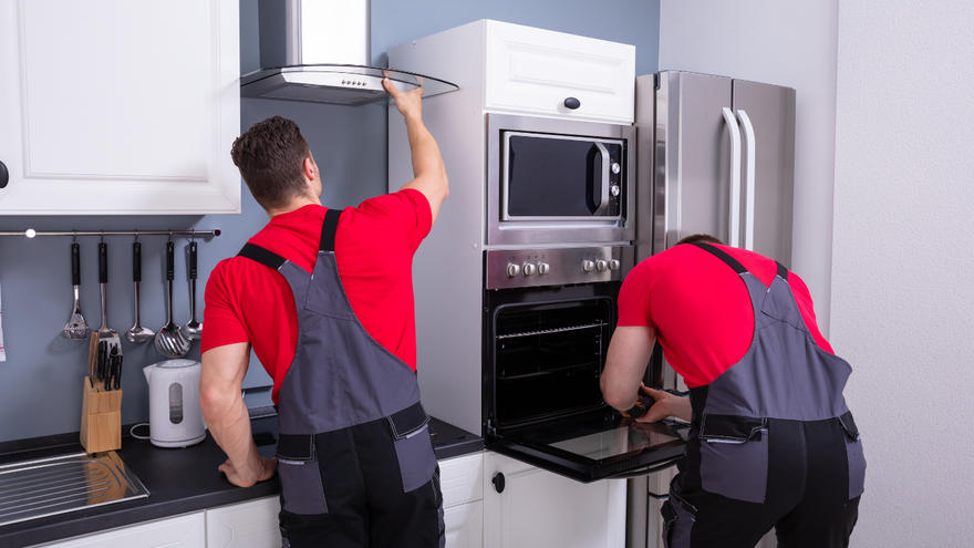 Two technicians in red shirts and grey overalls work on kitchen appliances, including a range hood and wall-mounted oven.