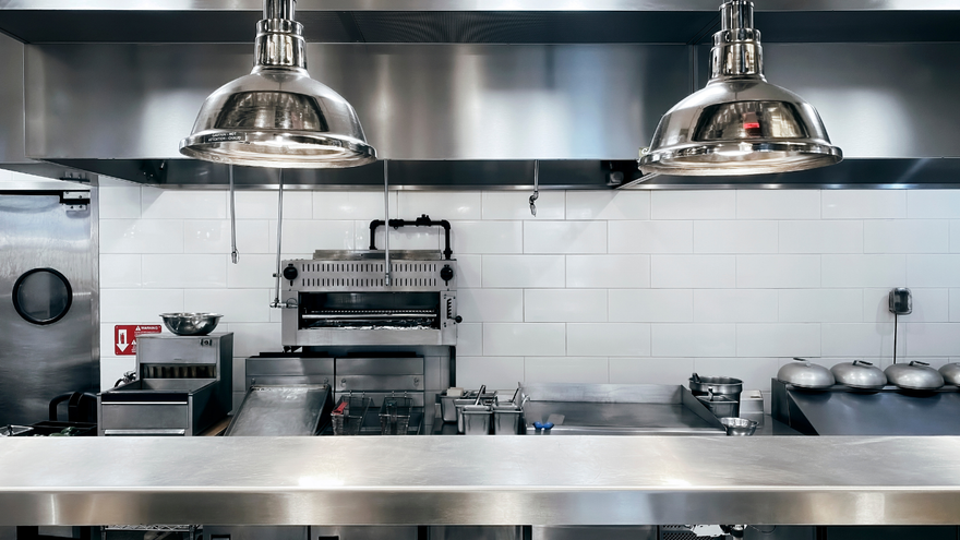 A stainless steel commercial kitchen counter with two hanging pendant lights, cooking equipment, and a white tiled wall.