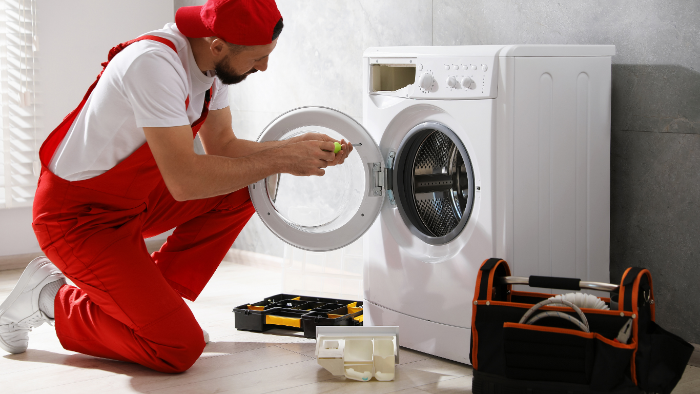 A technician in red work clothes kneels while repairing the open door of a white washing machine.
