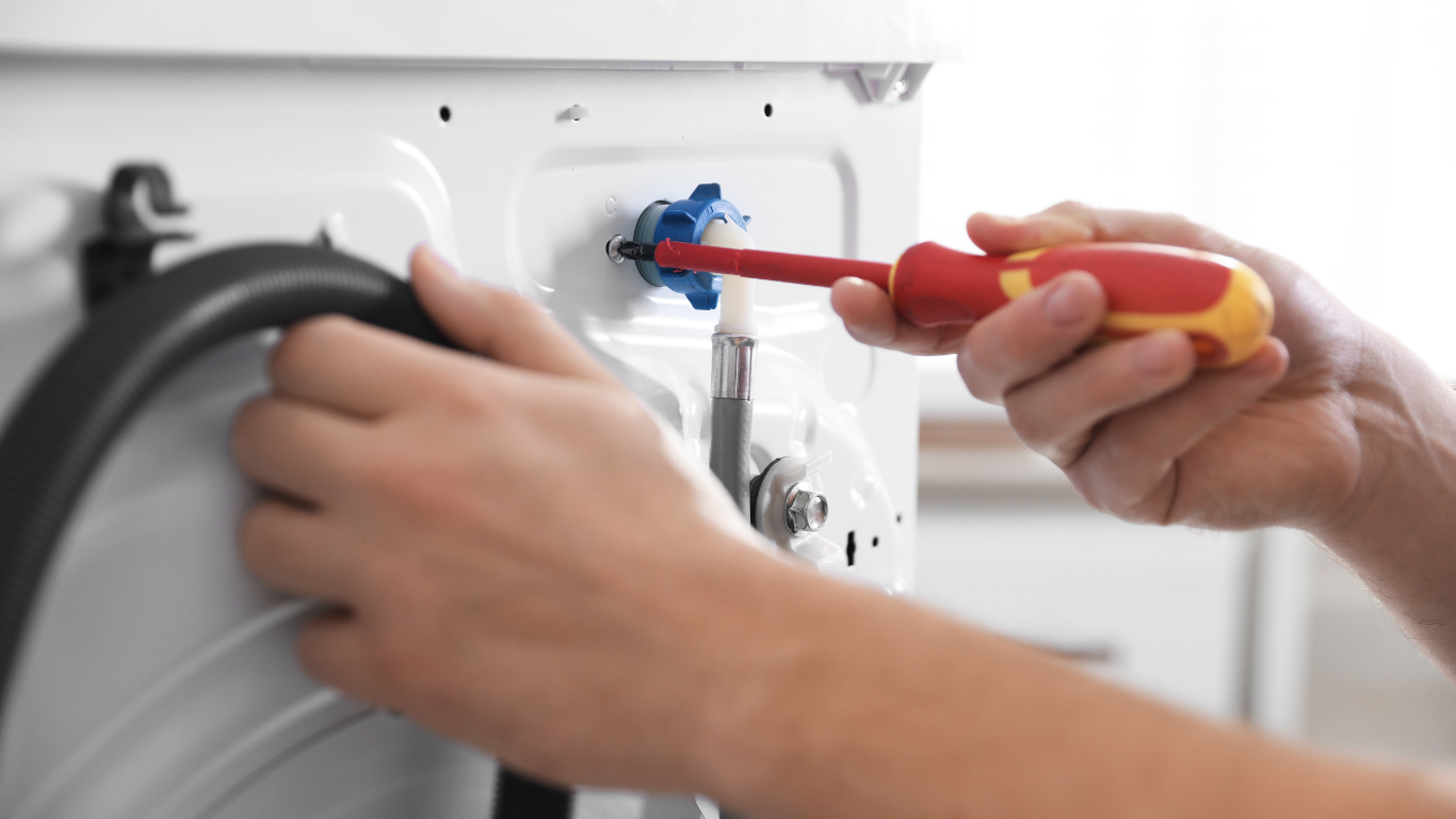 A person uses a red and yellow screwdriver to adjust a blue inlet valve on the back of a white washing machine.