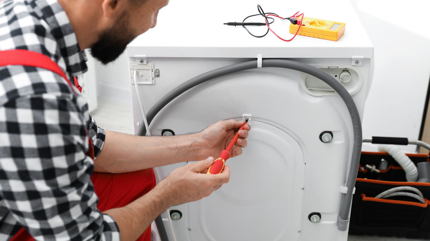 A technician in a plaid shirt uses a screwdriver on the back of a washing machine next to a yellow multimeter.
