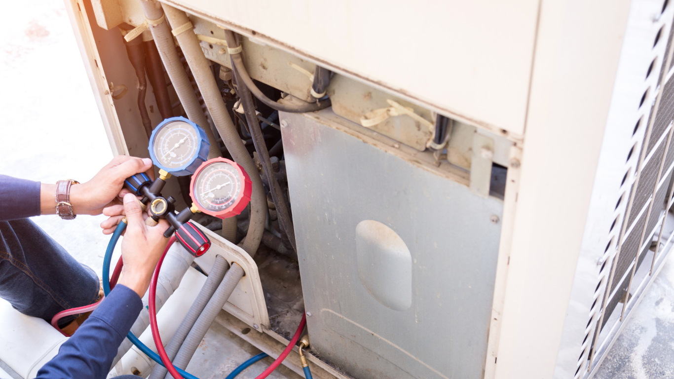 A technician holds a manifold gauge set to check refrigerant pressure on an outdoor air conditioning unit.
