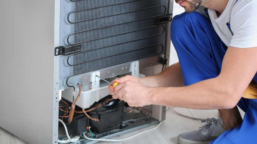 A technician in blue workwear uses a screwdriver to repair the back components of a refrigerator.