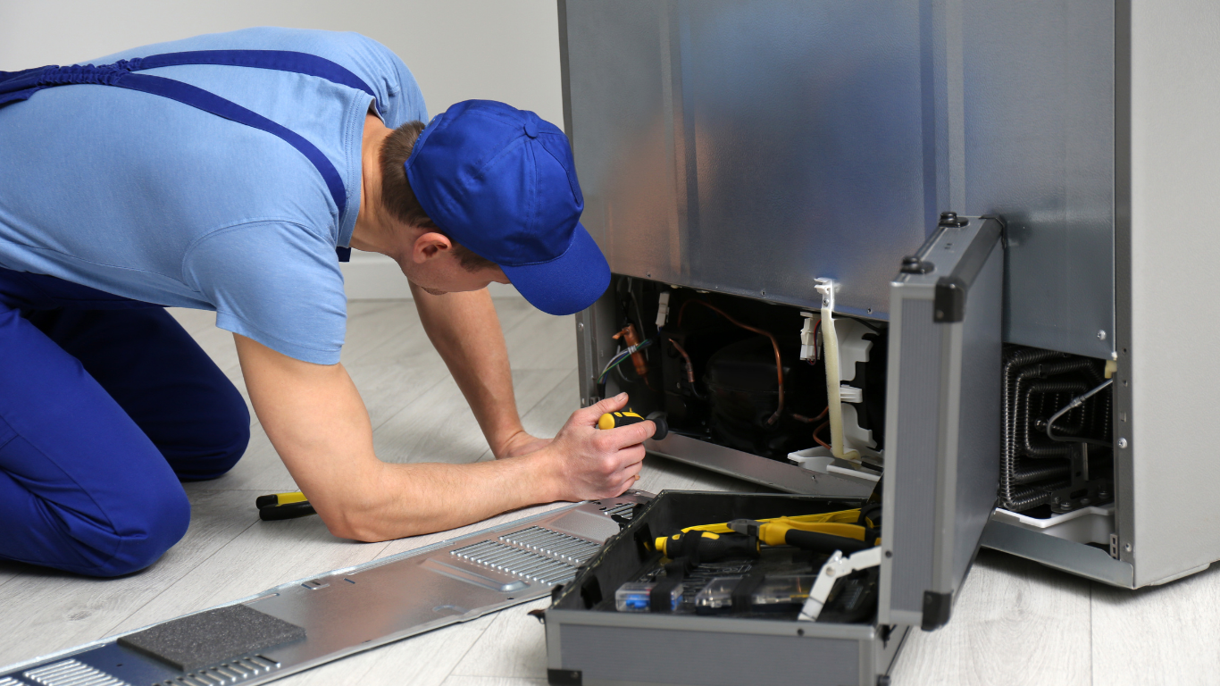 A technician wearing a blue uniform kneels on a floor, repairing the open back panel of a refrigerator with tools nearby.