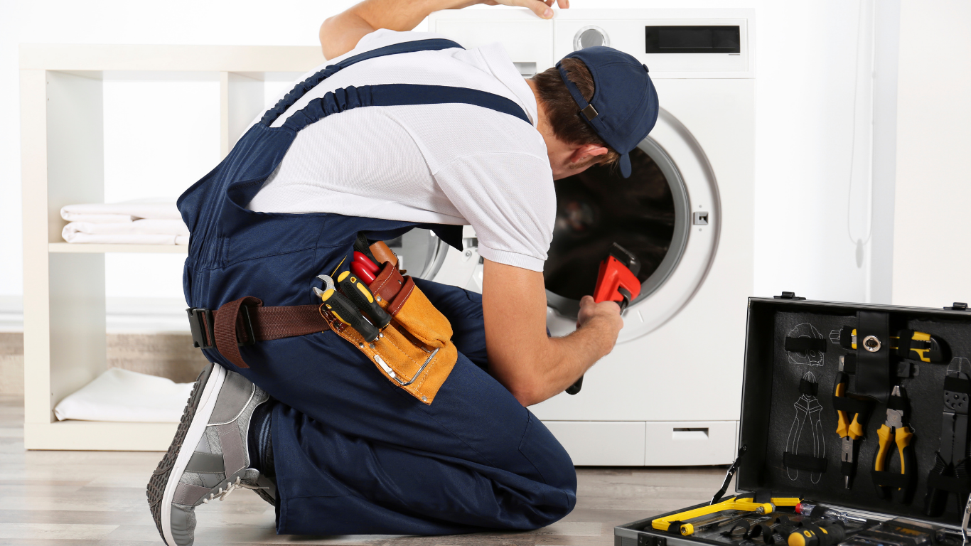 A service technician in blue overalls kneeling to repair a white washing machine, with a tool belt and tool case nearby.