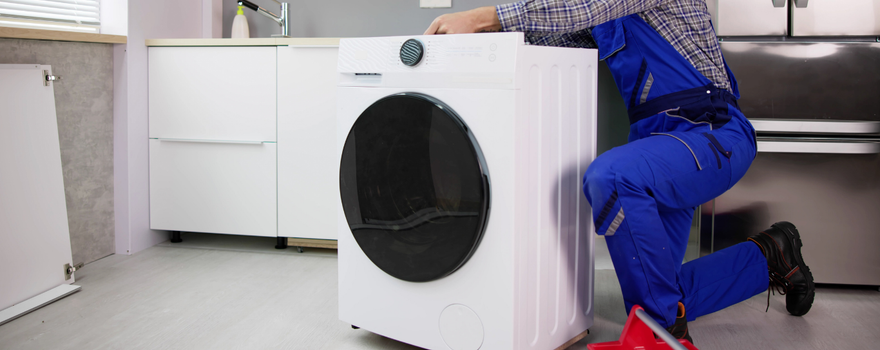 A worker in blue overalls kneeling and installing a white front-load washing machine in a kitchen.