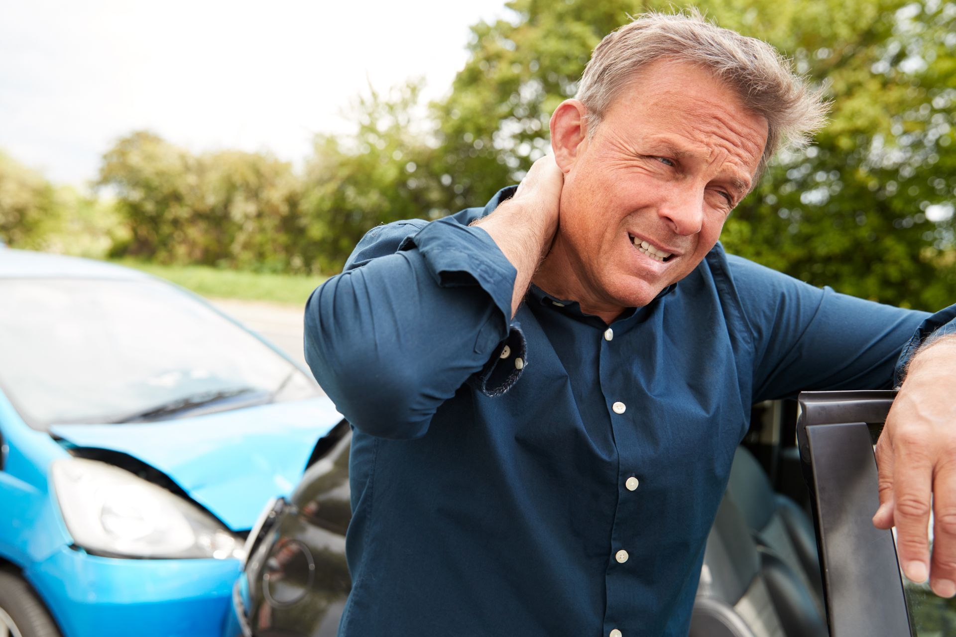 A man is standing in front of a blue car with his hand on his painful neck.