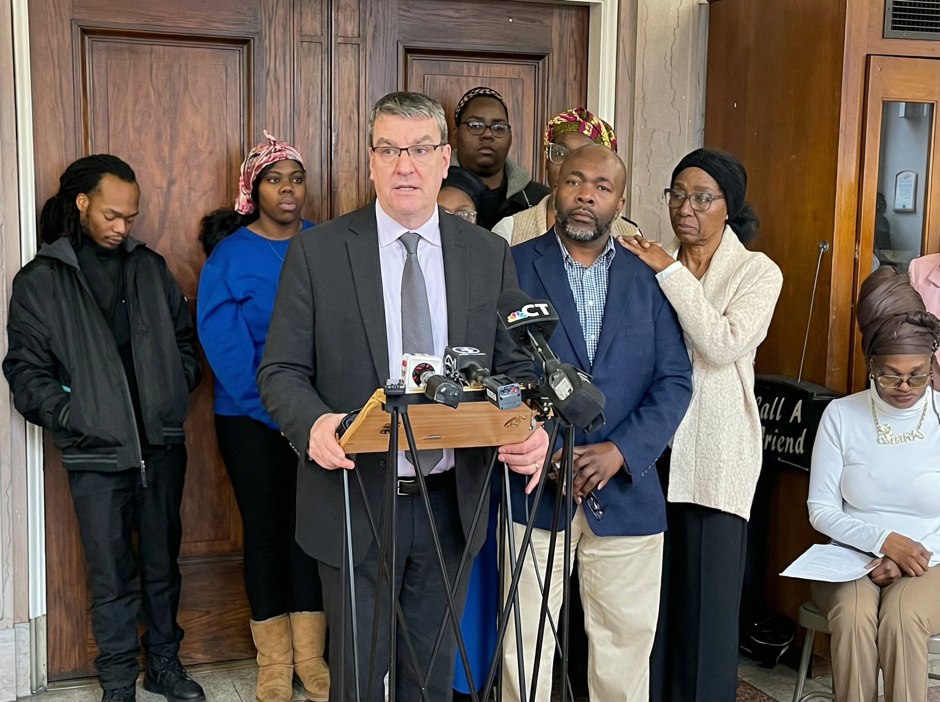 Man speaking at a podium, surrounded by people outside a building, media microphones present.