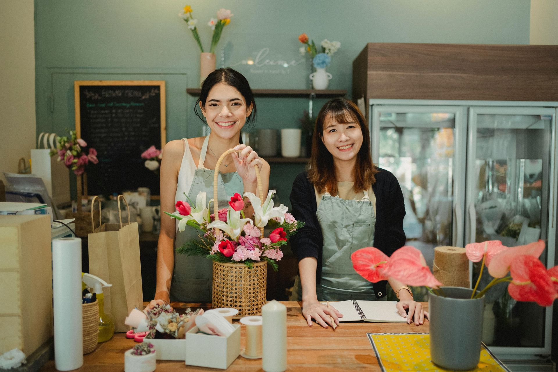 Two women smiling behind the counter of a flower shop, floral arrangements displayed.