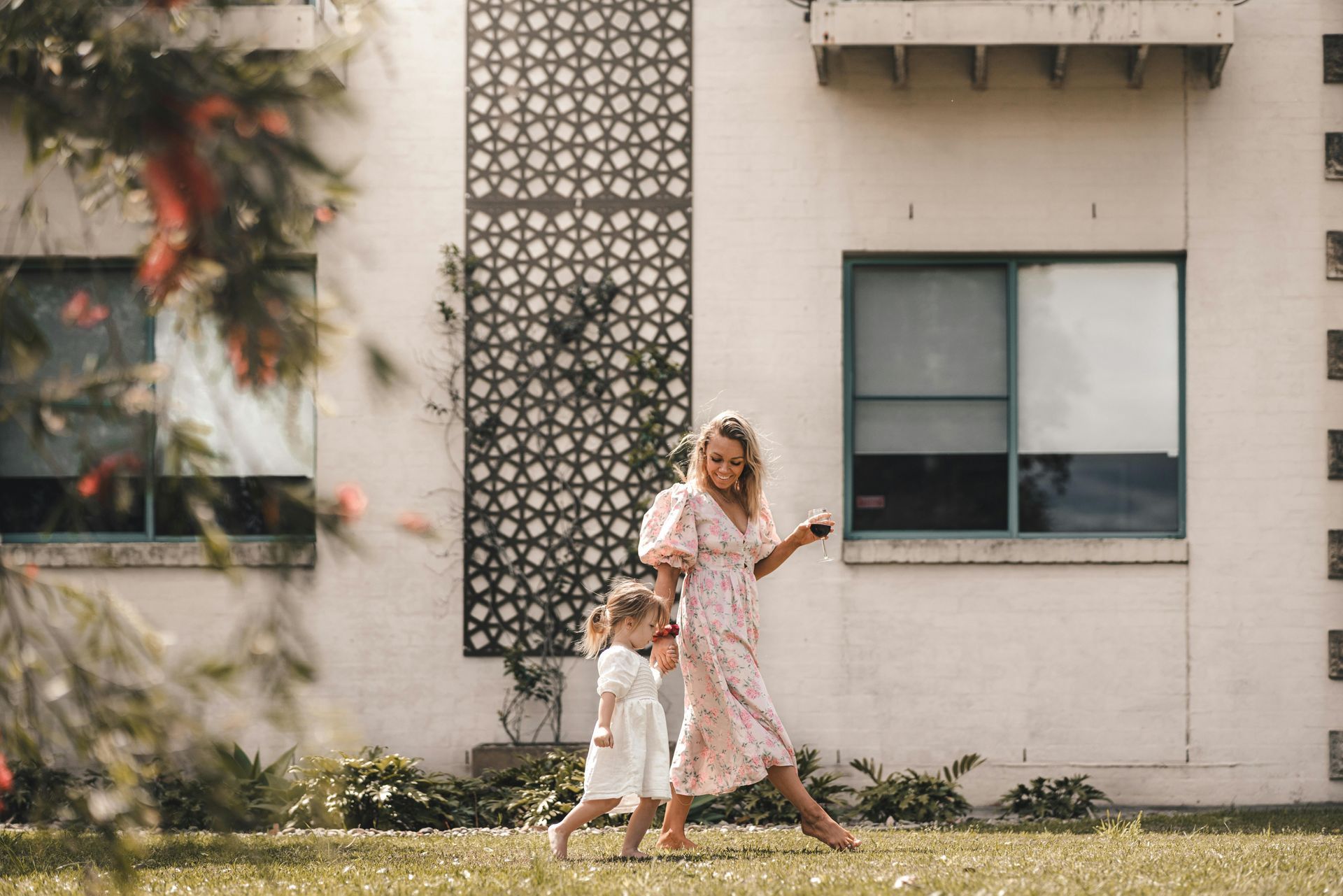 Woman in pink dress walking with child in white dress on lawn. White building background.