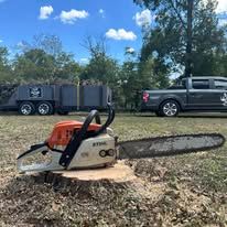 Orange chainsaw on a cut stump with a pickup truck and trailer in the background under a blue sky