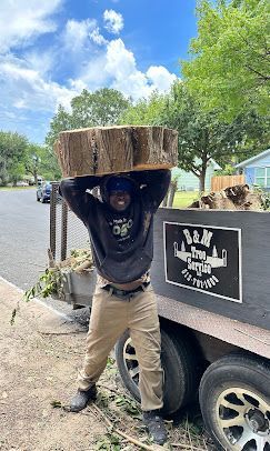 Person lifting a large log beside a trailer on a sunny street, trees and blue sky in the background